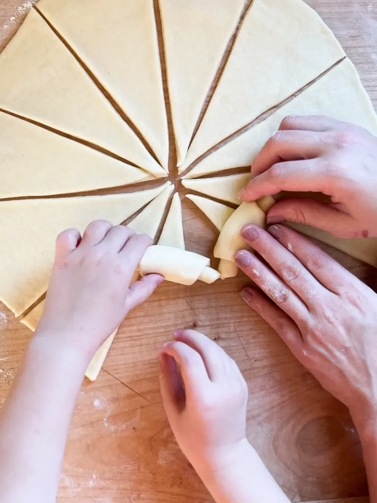 Two sets of hands, one mother and one child, rolling up the wedges into crescent shapes.