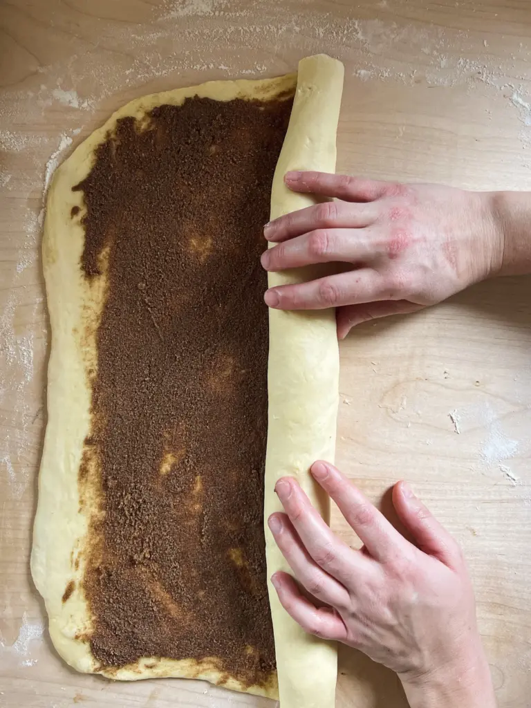 Soft sourdough cinnamon roll dough being rolled up tightly with a buttery cinnamon-sugar filling, ready to be sliced.