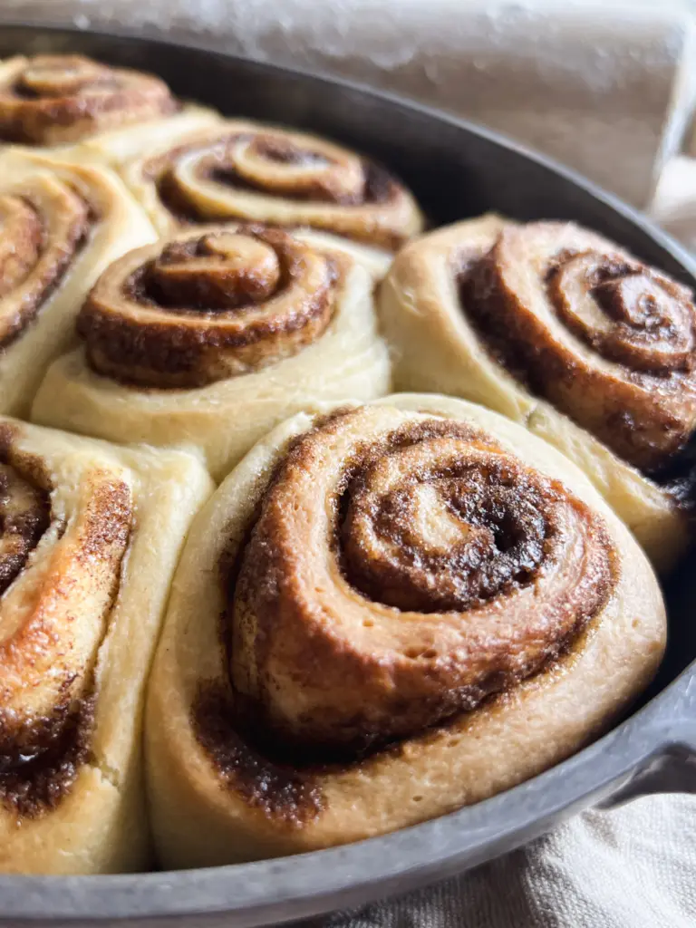 Close-up of freshly baked sourdough cinnamon rolls in a pan, golden brown with visible cinnamon swirls, just before being frosted.