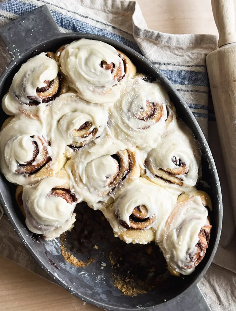 A pan of freshly baked sourdough cinnamon rolls generously frosted with cream cheese icing, sitting on a rustic countertop next to a wooden rolling pin.