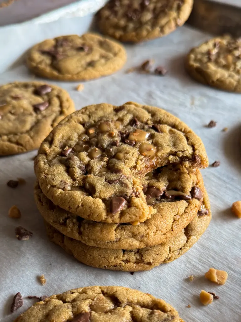 Stack of chewy Heath Bar Toffee Cookies on parchment paper, with melted toffee and chocolate pieces visible. The top cookie has a bite taken out, revealing its soft, gooey center.
