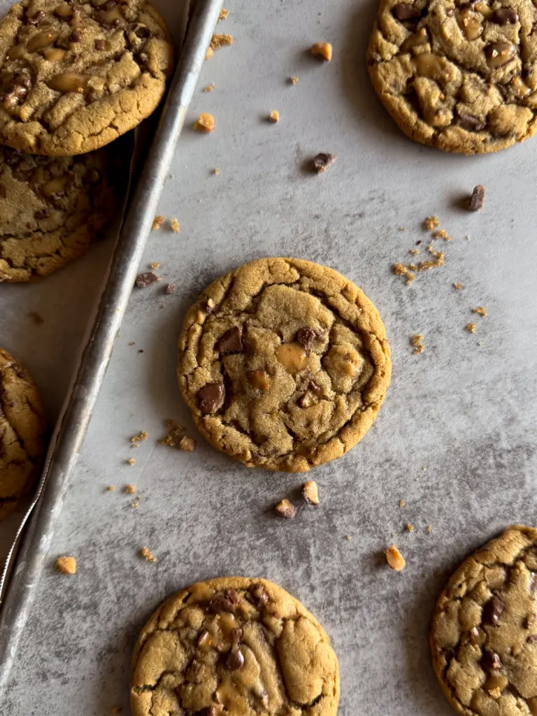 Close-up of a single Heath Bar Toffee Cookie on a baking sheet, surrounded by scattered toffee bits and chocolate chunks. Cookie is golden brown with a crackly top and visible toffee pieces.