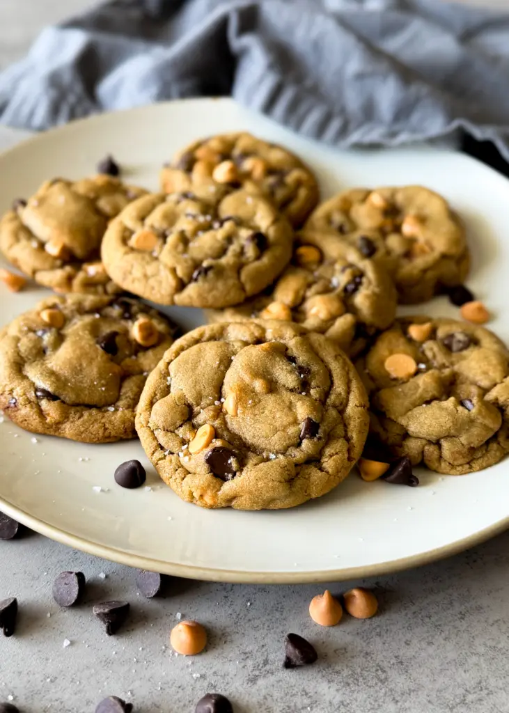 Plate of sea salt sprinkled chocolate chip butterscotch cookies, surrounded by scattered chocolate and butterscotch chips.