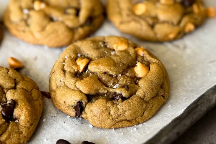 Chewy chocolate chip butterscotch cookies sprinkled with flakey sea salt on a parchment-lined baking sheet.